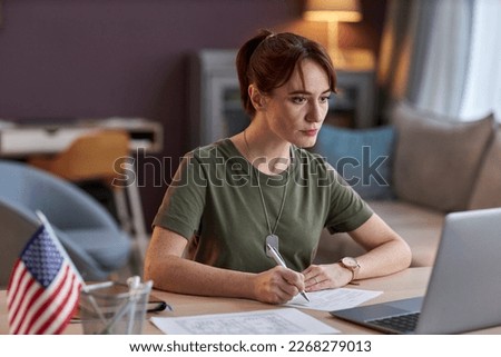 Portrait of young military woman looking at laptop screen and taking notes in online meeting