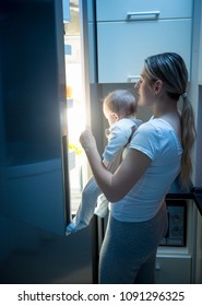 Portrait Of Young Mother Holding Baby Opens Refrigerator To Find Some Food At Night