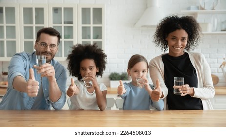 Portrait of young multiracial family with two children drink clean still mineral water together in kitchen. Smiling multiethnic mom and dad with diverse daughters show thumbs up recommend hydration. - Powered by Shutterstock