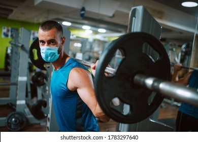 Portrait Of A Young Muscular Man With A Medical Face Mask Lifting Weights In The Gym. COVID 19 Coronavirus Protection