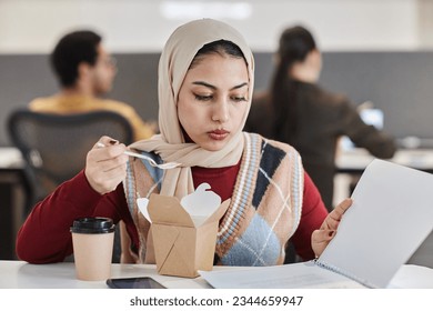 Portrait of young Muslim businesswoman eating takeout food at office workplace and reading documents, copy space - Powered by Shutterstock