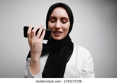 Portrait of young muslim woman sending audio voice message on cellphone talking to assistant. Wearing black hijab.