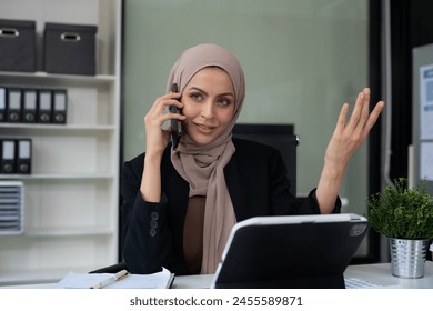 Portrait of young muslim woman talking mobile phone and working on laptop and papers on table in her home office. - Powered by Shutterstock