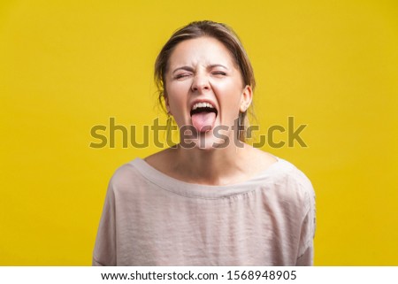 Portrait of young naughty woman with blonde hair in casual beige blouse standing with closed eyes and tongue out, teasing grimace, facial expression. indoor studio shot isolated on yellow background