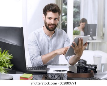 Portrait of young newspaper editor sitting at workplace and holding his hands digital tablet while working online. 