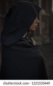 Portrait of young nun in black cassock and hooded cloak with cross in her hands. Side view.