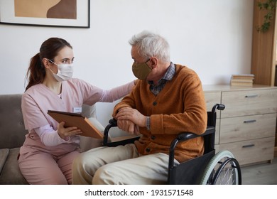 Portrait of young nurse assisting senior man in wheelchair using digital tablet at retirement home, both wearing masks, copy space - Powered by Shutterstock