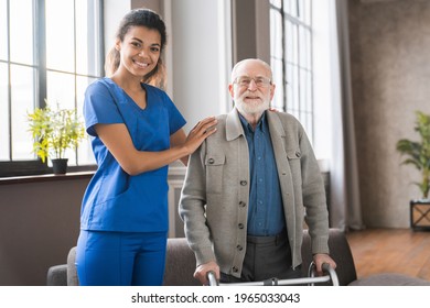 Portrait Of Young Nurse Helping Senior Man To Walk Using Walker Equipment In The Bedroom. Nurse With Old Elderly Grandfather Using Walker In Rehab Center.