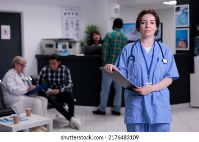 Portrait Of Young Nurse With Stethoscope In Busy Private Hospital Reception Holding Clipboard Looking At Camera. Professional Healthcare Worker Posing Confident Waiting For Next Appointment.