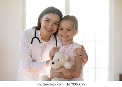 Portrait Of Young Nurse Wearing White Coat, Embracing Shoulders Of Happy Little Preschool Patient. Smiling Small Girl Holding Favorite Toy, Posing For Photo With Attractive General Practitioner.