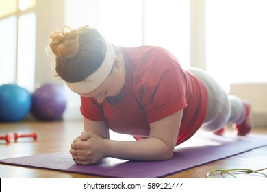 Portrait Of Young Obese Woman Working Out On Yoga Mat In Sunlit Fitness Studio: Holding Plank Exercise With Effort To Lose Weight