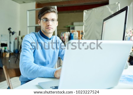 Portrait of young office worker wearing glasses using PC sitting at desk in modern office, copy space