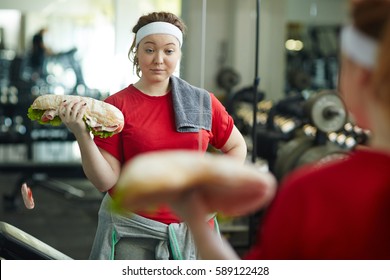 Portrait Of Young Overweight Woman Doing Weight Exercises With Huge Fat Sandwich While Working Out In Gym, Struggling To Keep Fit