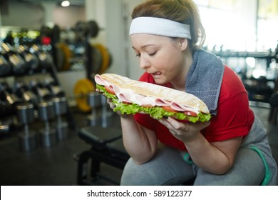 Portrait Of Young Overweight Woman Licking Her Lips Wanting To Eat Huge Fat Sandwich While Working Out In Gym, Struggling To Keep Fit