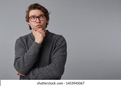 Portrait of young pensive man with glasses wears in gray sweater, stands over gray background with copy space on the right side, touches chin and looks up. - Shutterstock ID 1453945067