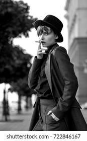 Portrait of a young pensive woman in man's suit and hat smoking a cigarette on a street, black and white