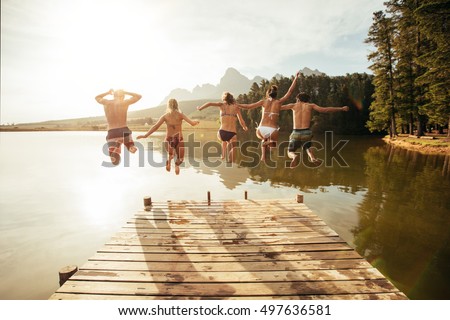 Portrait of young people jumping from pier into lake together. Friends jumping off the jetty at the lake on a sunny day.