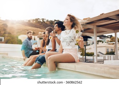 Portrait of young people sitting on the edge of the swimming pool with their feet in water. Friends relaxing by the pool during a party. - Powered by Shutterstock