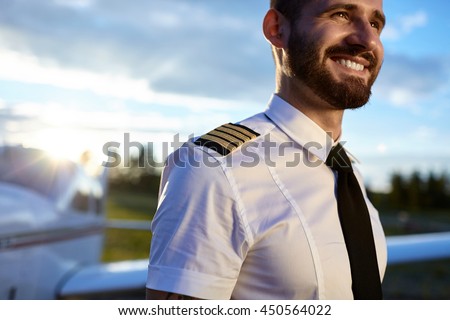 Portrait of young pilot in uniform posing with a happy toothy smile with air craft on the background. Backlit sunny photo