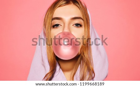 Portrait of young playful blonde girl dressed in blue hoodie standing isolated over pink background while blowing bubble with chewing gum. Woman looking at camera