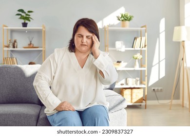 Portrait of a young plump overweight woman sitting on the couch in the living room at home and suffering from headache. Sick fat girl feel stressed, migraine or depression. Healthcare concept. - Powered by Shutterstock