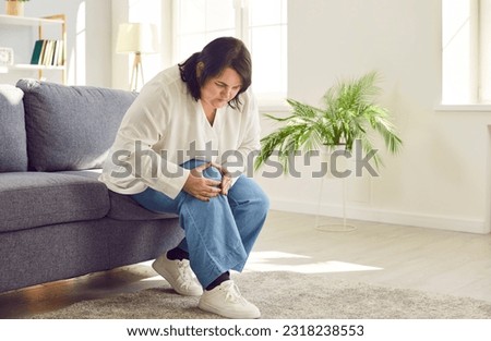 Portrait of young plus size overweight woman holding a knee suffering from osteoarthritis or arthritis sitting on a sofa in the living room at home. Obesity, chronic disease, health care concept.