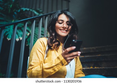 Portrait of young positive girl using modern smartphone device typing text message in social media while sitting on stairs outdoor, happy female student spending free time in break enjoying music - Powered by Shutterstock