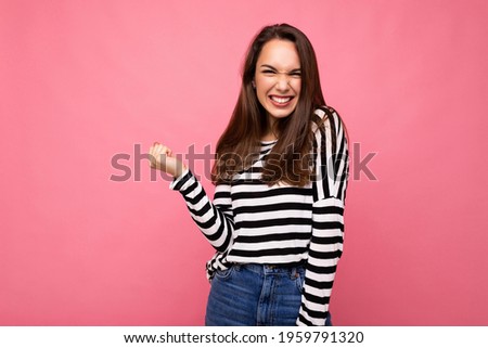 Portrait of young positive happy attractive brunette woman with sincere emotions in casual striped pullover isolated on pink background with free space and celebrating winning shouting yeah