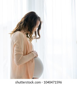 Portrait of young pregnant attractive woman, standing by the window, dressed in casual clothing, day before due date