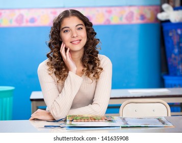 Portrait Of Young Preschool Teacher With Hand On Chin Sitting At Desk In Classroom