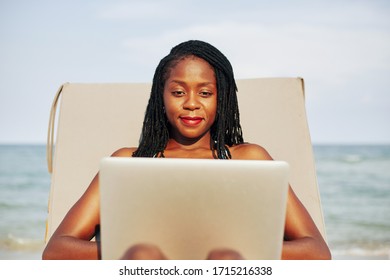 Portrait Of Young Pretty Black Woman Relaxing On Chaise-lounge And Working On Laptop On Beach