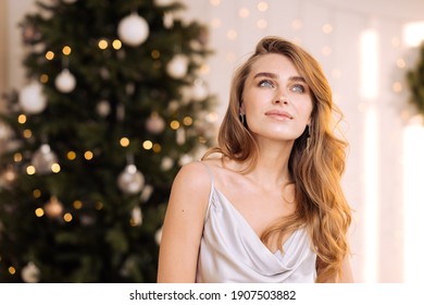 Portrait Of A Young Pretty And Dreamy Blonde Woman With A Silver Dress Against The Backdrop Of The Christmas Tree. Cozy Home Interior.