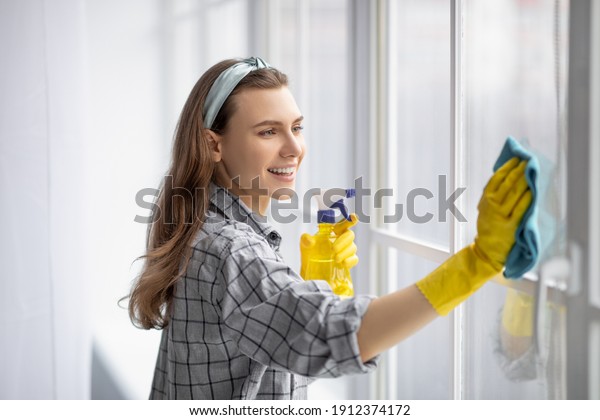 Portrait of young pretty lady in rubber gloves\
cleaning window with detergent at home. Lovely millennial housewife\
keeping her home tidy, happily performing domestic chores, enjoying\
housework