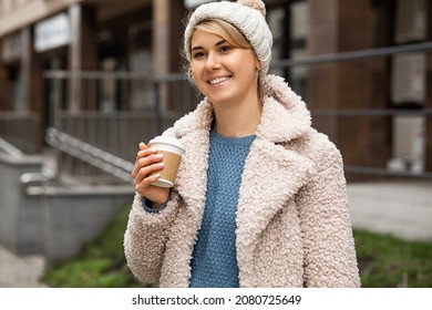 Portrait Of Young Pretty Woman 28 Years Holds Disposable Recyclable Cup With Hot Drink Of Coffee Or Tea In Hand. Smiling, Wearing Hat, Fur Coat. Street Style. Great Mood, Happy People.