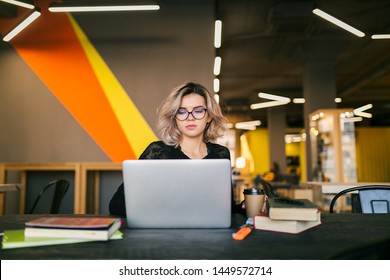 Portrait Of Young Pretty Woman Sitting At Table In Black Shirt Working On Laptop In Co-working Office, Wearing Glasses