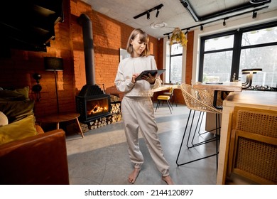 Portrait Of A Young Pretty Woman Standing With A Digital Tablet In Cozy Living Room. Wide View On Interior In Loft Style With Burning Fireplace On Background