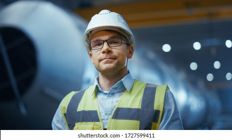 Portrait of Young Professional Heavy Industry Engineer / Worker Wearing Safety Vest and Hardhat Smiling on Camera. In the Background Unfocused Large Industrial Factory where Welding Sparks Flying.
