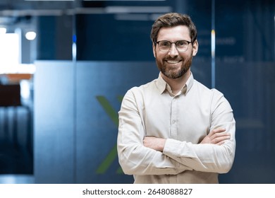 Portrait of a young programmer and engineer standing in the office with his arms crossed on his chest and smiling at the camera.