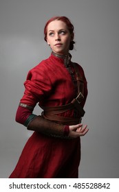 Portrait Of A Young  Red Haired  Female Warrior,  Wearing A Red Medieval Tunic And Leather Armour.  Grey Background.