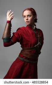 Portrait Of A Young  Red Haired  Female Warrior,  Wearing A Red Medieval Tunic And Leather Armour.  Grey Background.