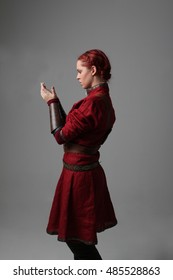 Portrait Of A Young  Red Haired  Female Warrior,  Wearing A Red Medieval Tunic And Leather Armour.  Grey Background.