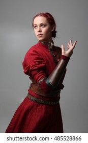 Portrait Of A Young  Red Haired  Female Warrior,  Wearing A Red Medieval Tunic And Leather Armour.  Grey Background.