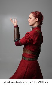 Portrait Of A Young  Red Haired  Female Warrior,  Wearing A Red Medieval Tunic And Leather Armour.  Grey Background.