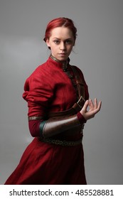 Portrait Of A Young  Red Haired  Female Warrior,  Wearing A Red Medieval Tunic And Leather Armour.  Grey Background.