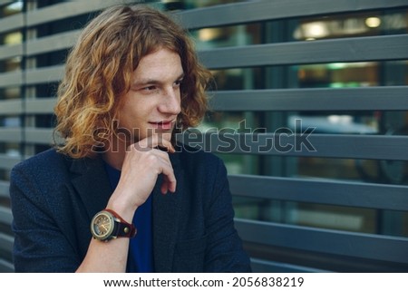 Portrait of young red haired, reddish man with curly hair, gray jacket, blue t-shirt and vintage watch over gray urban futuristic background, touching thoughtful his mouth with arm.