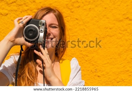 portrait of young red haired woman taking a picture holding a camera on colorful background wall in summer. happy and joyful caucasian girl smiling at camera making a photo. Copy space