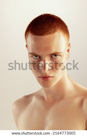 Portrait of young red-haired man with freckled face posing, seriously looking at camera isolated over grey studio background. Concept of men's health, lifestyle, beauty, body and skin care, fashion