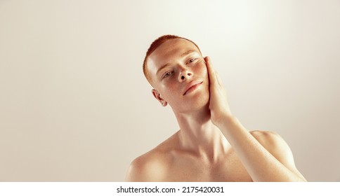Portrait of young red-haired man with freckles touching cheeks, posing isolated over grey studio background. Shaving lotion. Concept of men's health, lifestyle, beauty, body and skin care