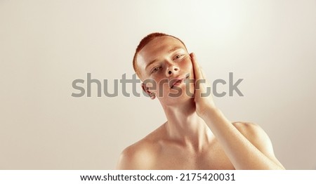 Portrait of young red-haired man with freckles touching cheeks, posing isolated over grey studio background. Shaving lotion. Concept of men's health, lifestyle, beauty, body and skin care
