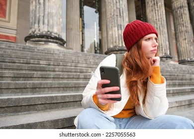 Portrait of young redhead woman with complicated face, sits on street stairs in red hat, holds smartphone and frowns thoughtful, feels uneasy. - Shutterstock ID 2257055563